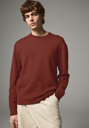 Bordeauxfarbenes Strickpullover mit Rundhalsausschnitt, gerippten Bündchen und lässiger Passform. Kombiniert mit hellbeigen Hosen, die eine glatte Textur aufweisen.