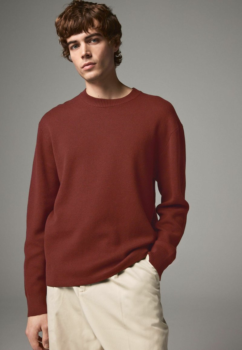 Maglione a maglia bordeaux con scollo rotondo, polsini a coste e vestibilità rilassata. Abbinato a pantaloni beige chiaro, che mostrano una texture liscia.