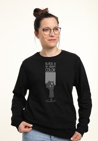Donna con occhiali e chignon, che indossa una felpa nera con la scritta "Il nero è il mio colore felice" e un design di silhouette che tiene un ombrello.