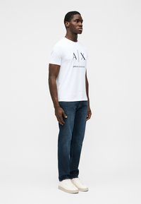 Tricou alb cu mâneci scurte, având un logo „A/X” negru îndrăzneț și textul „ARMANI EXCHANGE”, asortat cu blugi de culoare albastru închis și adidași albi.