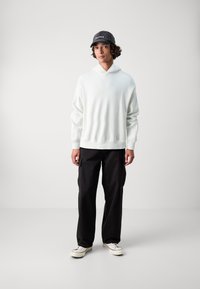 Λευκή φούτερ μπλούζα με κουκούλα και πλεκτές μανσέτες, συνδυασμένη με μαύρα cargo παντελόνια και λευκά αθλητικά παπούτσια. Το μοντέλο φοράει ένα σκούρο καπέλο με κείμενο.