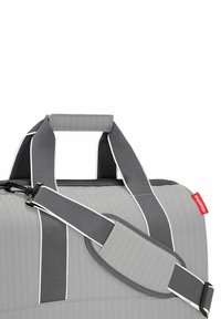 Grauem Duffelbag mit Fischgrätenmuster, ausgestattet mit einem gepolsterten Griff, verstellbarem Riemen und weißen Akzenten. Markenlabel in Rot an der Seite.