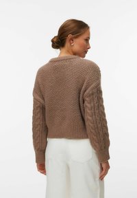 Femme avec les cheveux en chignon portant un pull beige en maille épaisse avec des manches à motif torsadé et un pantalon blanc taille haute, vue de profil.