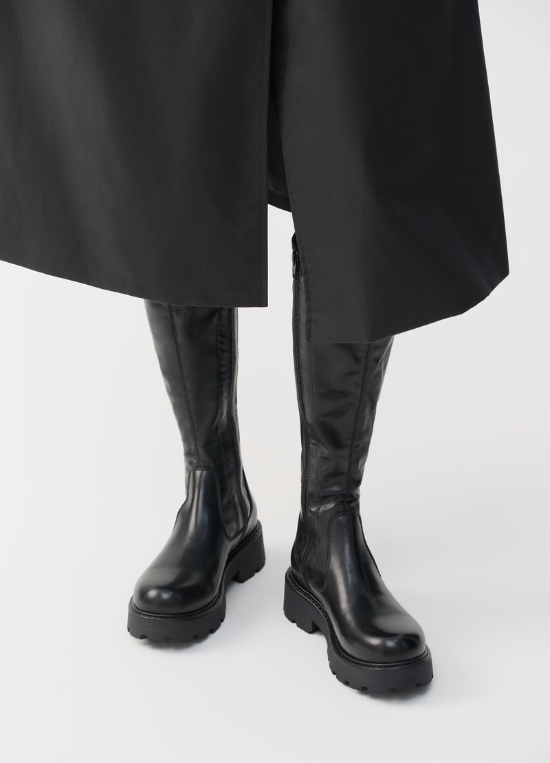 Bottes noires à hauteur de genoux avec une texture en cuir lisse, des semelles épaisses et des zips sur le côté pour un enfilage facile. Design élégant et fonctionnel.