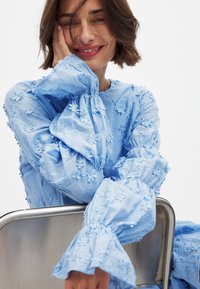 Svetlo modra vezenina bluzica z cvetličnimi vzorci, dolgi naborki, volančasti manšeti in okroglim izrezom, izdelana iz lahkega materiala.