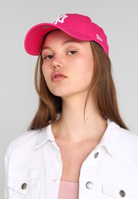 Rosa keps med vit logotyp, i bomullsmaterial, böjd skärm. Modell som bär en vit denimjacka över en ljusrosa topp, neutral bakgrund.