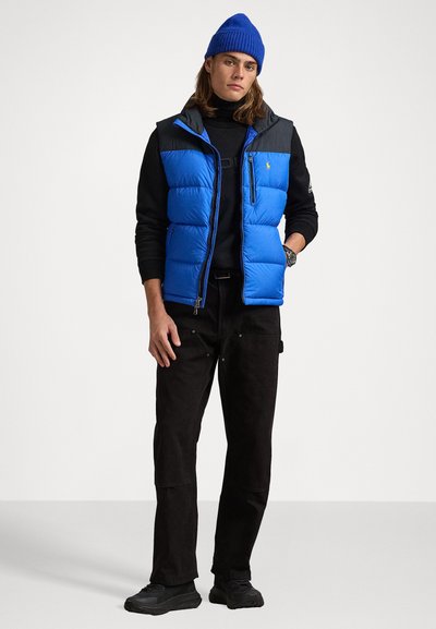 Zila pufota veste ar pelēkiem plecu akcentiem, ar rāvējslēdzi priekšpusē, valkāta virs melnas turtleneck krekla un melnām biksēm, apvienota ar melnām kurpēm un zilu cepuri.