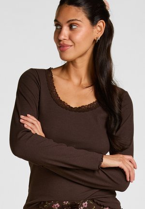 Vrouw met lang donker haar met gouden oorbellen in de vorm van hoepels en een bruine top met lange mouwen en kanten rand, armen gekruist, kijkt naar links.