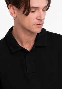 Musta polo-paitaa, jossa on teksturoitu kangas, klassinen kaulus ja pieni etupaneeli. Yksinkertainen ja minimalistinen muotoilu.