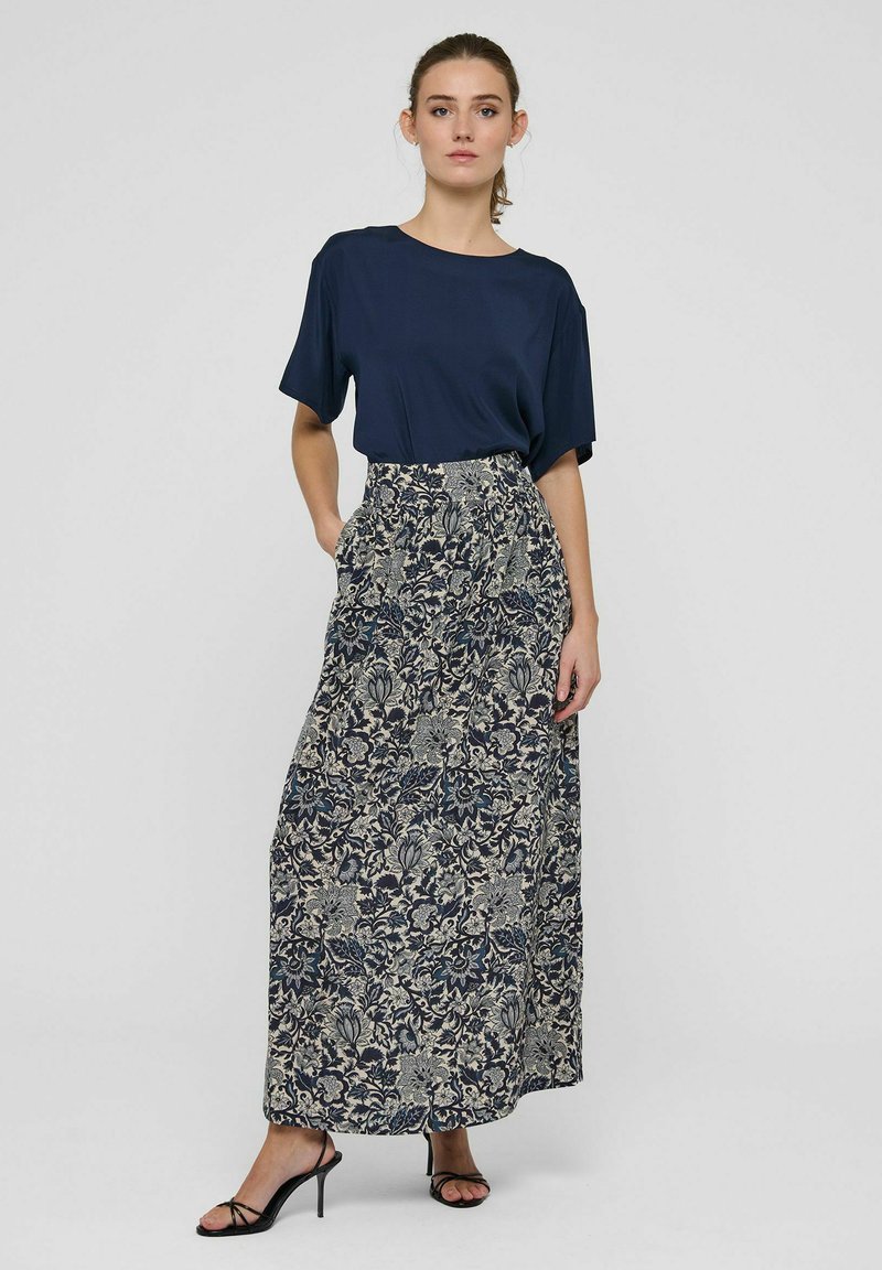 Femme portant un haut bleu marine à manches courtes et une longue jupe à motif floral avec des sandales noires à talons hauts sur un fond uni.