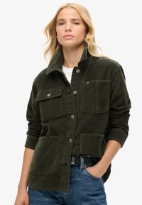 Giacca di velluto a coste verde scuro con collo a risvolto, due tasche sul petto e bottoni a pressione argentati. Abbinata a jeans e cintura nera.