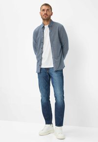 Herrenhemd in Blau mit Knöpfen und strukturiertem Stoff, getragen über einem weißen T-Shirt, kombiniert mit blauen Jeans und weißen Sneakern.