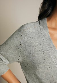 Lys grå strikket sweater med V-hals, der har mellemrumshuller langs skuldersømmene, og en spottet tekstur med farvede prikker.