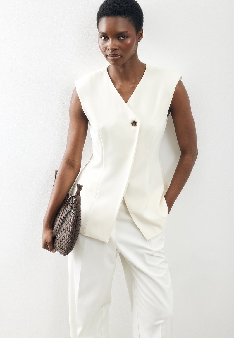 Woman wearing sleeveless white blazer and white pants, holding a brown woven clutch bag, posing against a plain white wall.