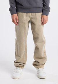 Beige henkareet, joissa on rento istuvuus, lahkeet käännettynä nilkoissa, yhdistettynä tummanharmaaseen collegepaitaan. Valkoiset tennarit, joissa on nauhakiinnitys.