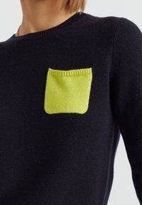 Maglione di colore blu navy con una vivace tasca verde lime. Il tessuto sembra morbido, con scollatura a costine e cuciture dettagliate sulla tasca.