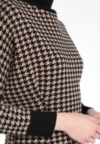 Pull en tricot avec un motif pied-de-poule noir et beige, avec un col et des poignets côtelés noirs.