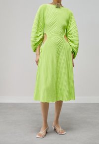 Abito plissettato verde lime con maniche a palloncino, ritagli laterali e dettagli floreali sul colletto. Indossato con décolleté bianchi con cinturini.