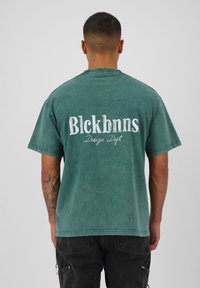 Groene, oversized katoenen T-shirt met korte mouwen. Heeft witte tekst "Blckbnns" en "Design Dept." op de achterkant, en een vervaagde textuur.