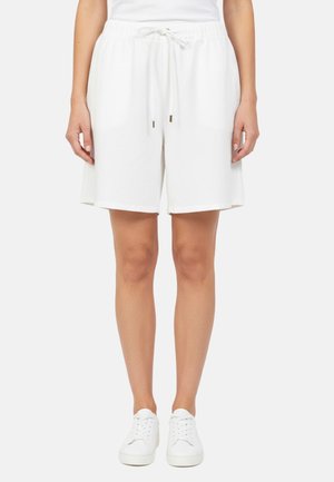 Donna che indossa pantaloncini bianchi fino al ginocchio con vita elasticizzata e coulisse, e sneakers bianche, in piedi davanti a uno sfondo semplice.