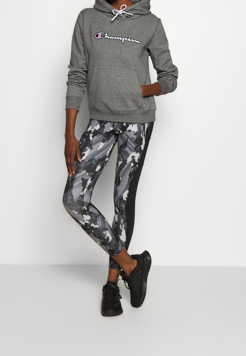 Personne portant un sweat à capuche gris Champion, des leggings à motifs noirs et blancs, et des chaussures de sport noires, debout sur fond blanc.