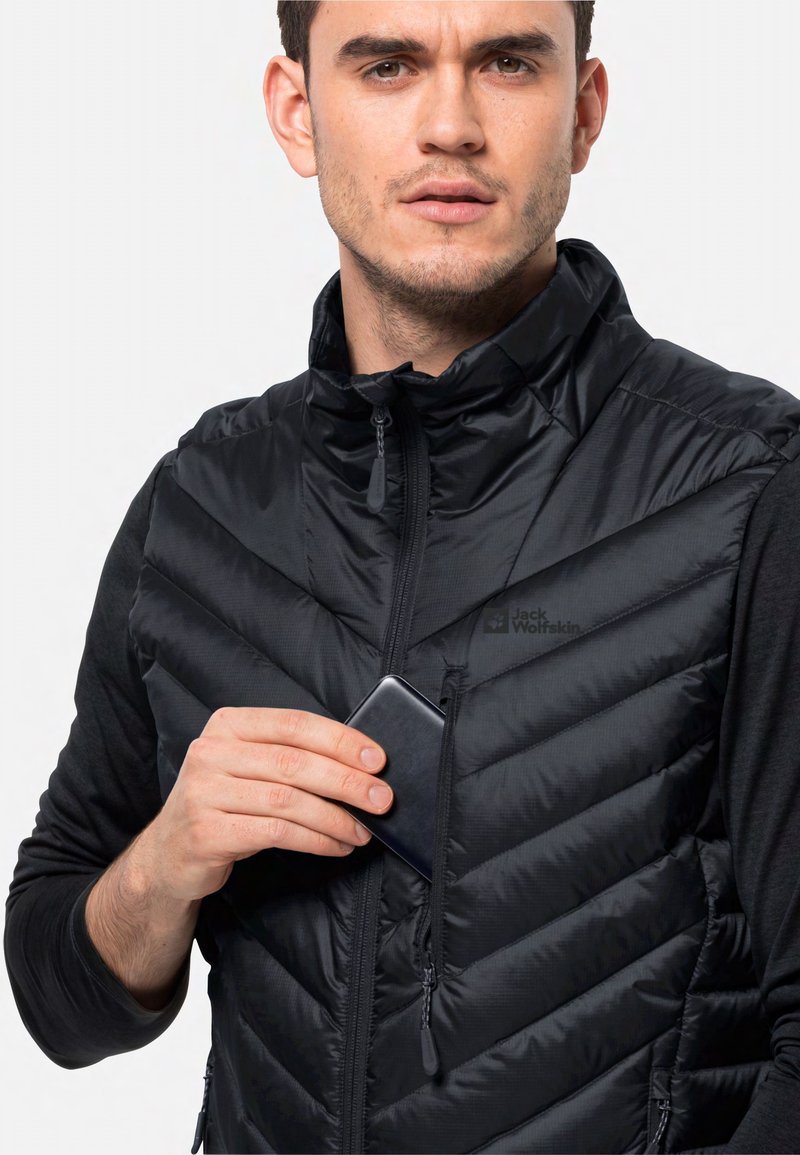 Schwarze gepolsterte Weste mit hohem Kragen und Zickzackmuster, ausgestattet mit einer mit Reißverschluss versehenen Brusttasche. Das Modell steckt ein Smartphone in die Tasche.