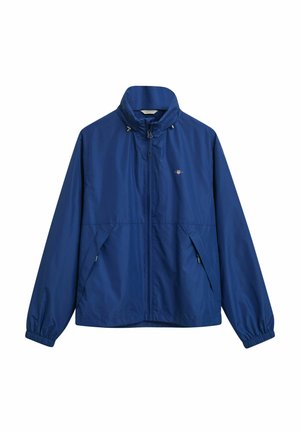 Blå lättviktsjacka med hög krage, elastiska ärmslut, blixtlåsfickor på sidorna och en subtil logotyp på bröstet. Slät textur.