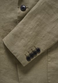 Manche d'un blazer beige avec quatre boutons noirs près du poignet et un bouton noir près du revers, montrant le détail du tissu texturé.