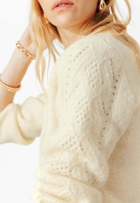 Pull en maille crème avec un motif torsadé texturé sur la manche, matériau doux et coupe légèrement oversize. Un éclat subtil ajoute de la dimension.