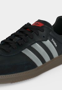 Μαύρο αθλητικό παπούτσι Adidas Samba με ασημένιες ρίγες, μαύρα κορδόνια, κόκκινη ετικέτα λογότυπου στη γλώσσα και σόλα από καουτσούκ.