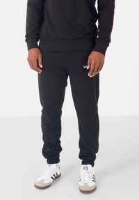 Pantaloni della tuta neri in morbido tessuto con taglio affusolato e polsini elasticizzati; presenta un logo bianco "SIX" sulla coscia sinistra.