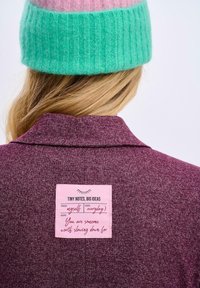 Bordowy żakiet z teksturowanej tkaniny. Różowa metka z tyłu zawiera odręczne notatki. Delikatnie zielona i różowa czapka widoczna powyżej.