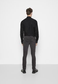 Hombre con camiseta de manga larga negra, jeans ajustados gris oscuro y zapatos negros de pie frente a una pared blanca, con la espalda visible.