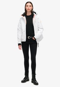 Witte waterdichte jas met een zwarte binnenkant, voorzien van oranje accenten. Gecombineerd met een gestructureerde zwarte sweater, zwarte jeans en veterschoenen.