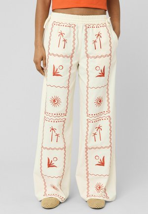Pantalon large couleur crème avec broderie orange sur le thème tropical, taille élastique et cordon, porté avec des chaussures tissées beige et un crop top orange.