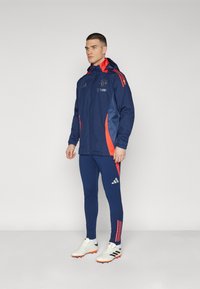 Chaqueta deportiva azul marino con acentos rojos y logotipo, combinada con leggings azul marino a juego. Presenta un cierre en la parte delantera y un diseño de tres rayas en las mangas.