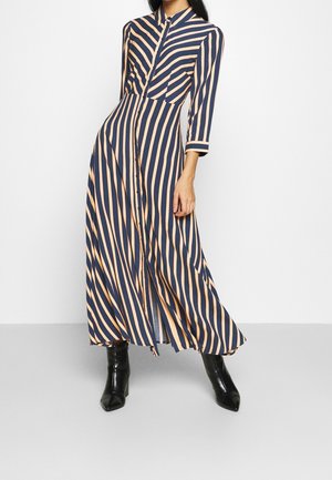 Kobieta w długiej sukience z pionowymi paskami w kolorze granatowo-beżowym, z rękawami do połowy przedramienia, nosząca czarne botki na obcasie.