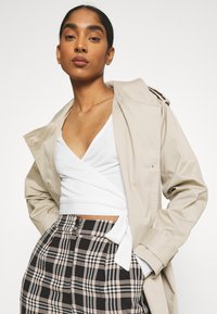 Capa bege sobre uma blusa branca envolvente, combinada com uma saia xadrez apresentando padrões em preto, branco e castanho. Texturas suaves.