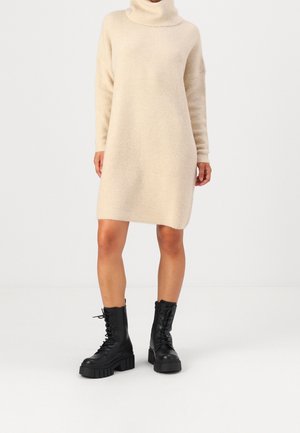 Beige geribbelde gebreide coltrui-jurk gedragen met zwarte chunky vetercombatlaarzen door een staand persoon.