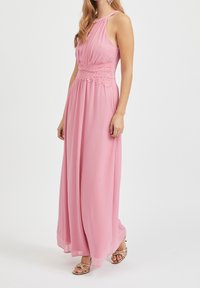 Robe longue rose clair avec un décolleté en forme de goutte, taille en dentelle froncée, tissu fluide et léger éclat, associée à des sandales dorées à brides.