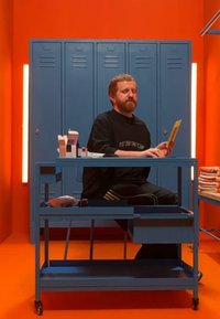 Carrito metálico azul con ruedas, cajón abierto y objetos encima. Fondo naranja brillante y taquillas azules detrás. Hombre sentado en el carrito.