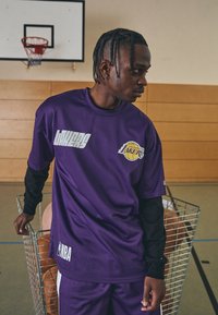 Camiseta de baloncesto de malla morada con el logo de los Lakers y el texto "NBA", acompañada de mangas largas negras, ubicada en un gimnasio con balones de baloncesto.