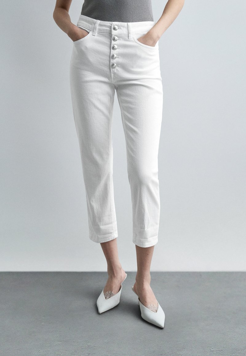 Γυναίκα που φοράει λευκό κοντό παντελόνι με πέντε κουμπιά και λευκά παπούτσια με μυτερή μύτη που μπαίνουν εύκολα, με τα χέρια στις τσέπες, όρθια σε γκρι δάπεδο.