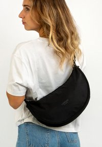 Borsa a spalla in tessuto nero con un design curvo, caratterizzata da una superficie liscia e un logo ben in evidenza. Indossata sopra una camicia bianca.