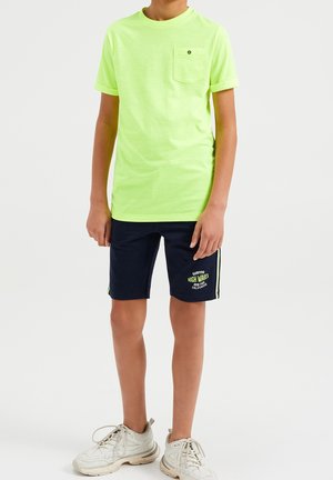 Neon groene katoenen T-shirt met een zakje, gecombineerd met marineblauwe shorts met witte tekst en gele strepen aan de zijkanten. Witte sneakers.