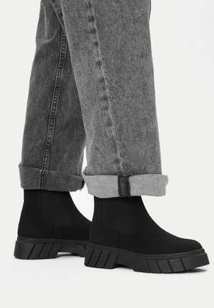 Czarne botki za kostkę wykonane z miękkiego materiału z masywną gumową podeszwą. Noszone z podwiniętymi szarymi spodniami jeansowymi, prezentujące prosty, elegancki design.