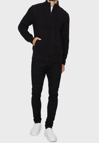 Pull noir zippé avec un design en maille torsadée texturée, col montant et deux poches latérales, associé à un pantalon noir et des baskets blanches.