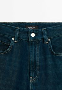 Ceinture de jeans en denim bleu foncé avec bouton argenté, passants pour ceinture et étiquette noire portant l'inscription « Massimo Dutti Made in Turkey ».