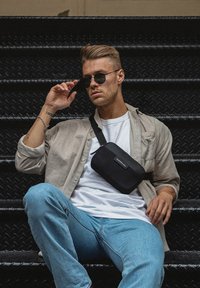Schwarze Umhängetasche aus glattem Material, mit Reißverschluss, Logodetail und verstellbarem Riemen. Getragen mit einem beigen Shirt und Jeans.