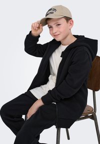 Sort zip-up hoodie og sweatpants, parret med en beige kasket med "New York City Athletics" i marineblå tekst. Sidder på en træstol.